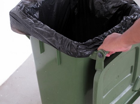 A close-up view of a green wheeled rubbish bin with a partially opened lid, revealing a black plastic bag liner filled with waste. The bin is positioned on a plain, grey concrete surface in an outdoor or driveway setting. A person's hand is visible gripping the handle on the side of the bin, suggesting it is being moved or prepared for waste collection. The scene is illuminated by natural daylight, highlighting the textured surface of the plastic liner and the smooth finish of the bin. This image is representative of waste removal services provided by Garden Clearance Camden, where lifting and handling of rubbish are common tasks, especially in garden or property clearance contexts within Camden or surrounding areas. The overall environment appears clean, functional, and typical of waste disposal activities in urban residential or commercial settings.