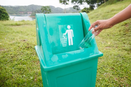 A person is seen disposing of transparent plastic bottles into a green recycling bin situated on a grassy area outdoors. The bin features a white pictogram of a figure discarding waste into a bin, alongside a waste collection symbol. The background includes a park-like environment with trees, a body of water, and a slightly elevated landscape, suggesting a natural setting in Camden or nearby. The scene is well-lit with natural daylight, and the surrounding grass appears lush and well-maintained. This image relates to waste management and recycling services offered by Garden Clearance Camden, emphasizing responsible disposal practices in a suburban or urban park environment within the postcode area. The person's hand is visible, and they are wearing a short-sleeved shirt, indicating mild weather conditions typical for the region.