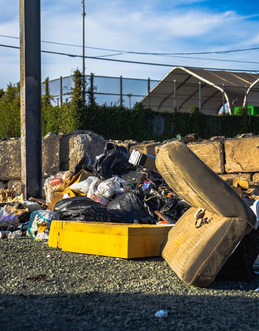 A close-up view of a pile of mixed household rubbish and waste materials accumulated on a paved surface in an outdoor area, featuring black plastic garbage bags, a yellow plastic container, and an old