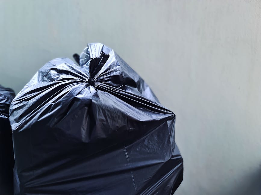 A close-up view of a pile of mixed household rubbish and waste materials accumulated on a paved surface in an outdoor area, featuring black plastic garbage bags, a yellow plastic container, and an old