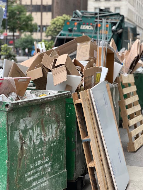 A street scene in an urban area showing a white garbage collection truck with an open rear hatch positioned on a cobblestone road, with visible signs of rust and wear on the metal surfaces. A worker d
